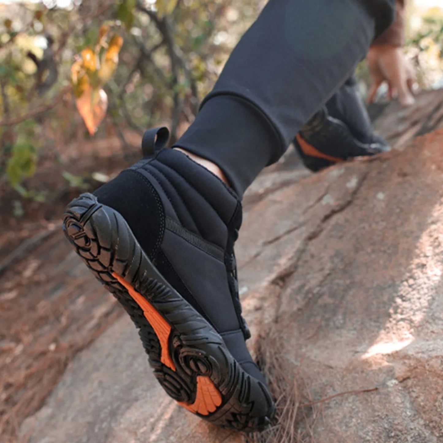 Black rugged hiking boots with orange traction worn by person walking on rock.