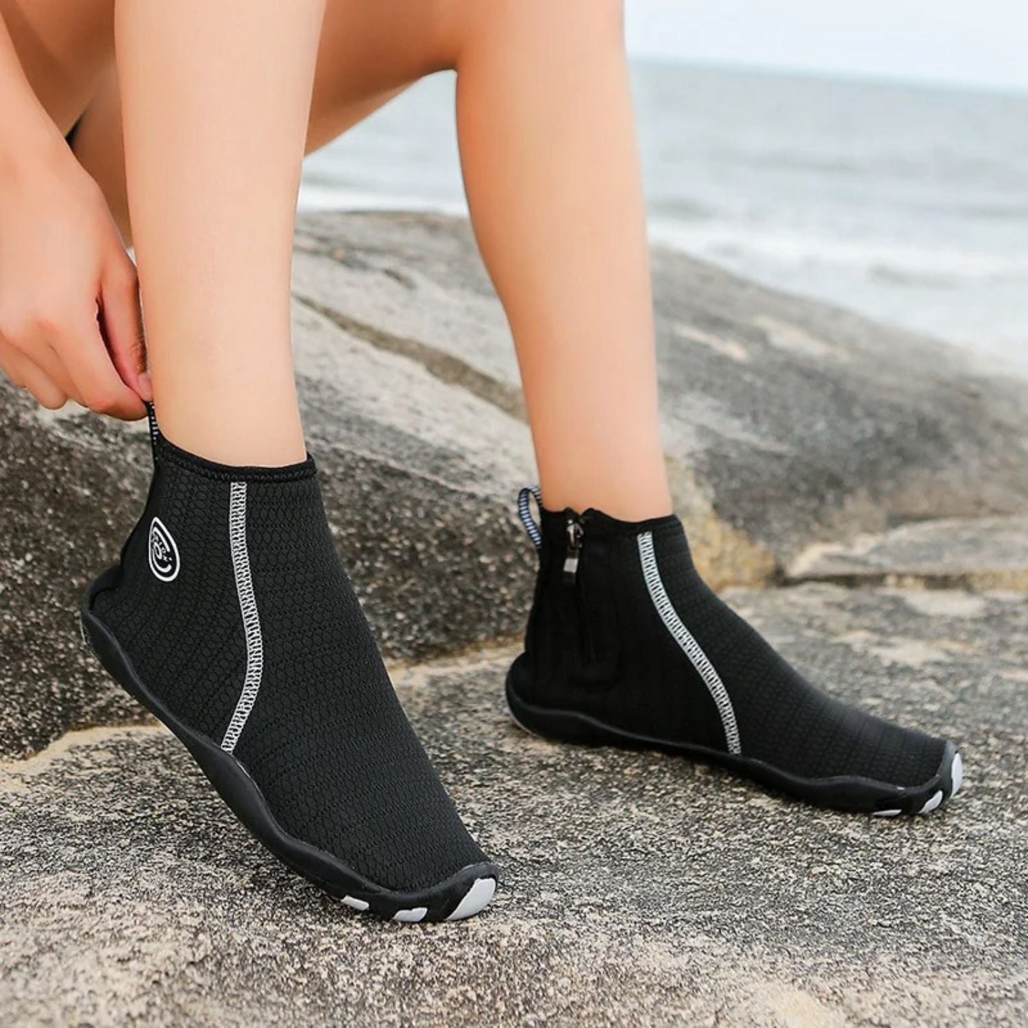 Black zip-up water shoes with white logo worn on beach, shown from the side.