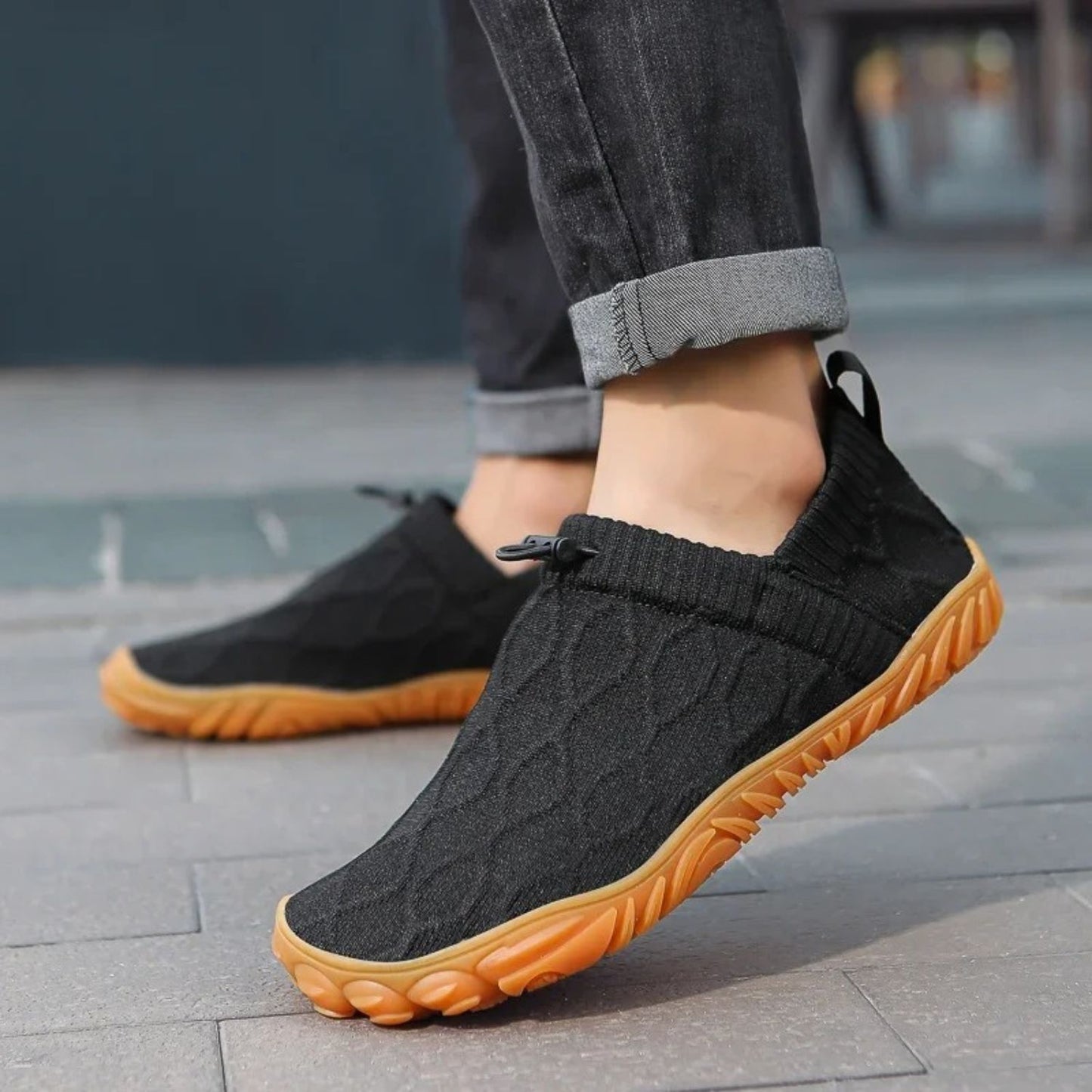 Person wearing black slip-on shoes with orange soles while walking on sidewalk.
