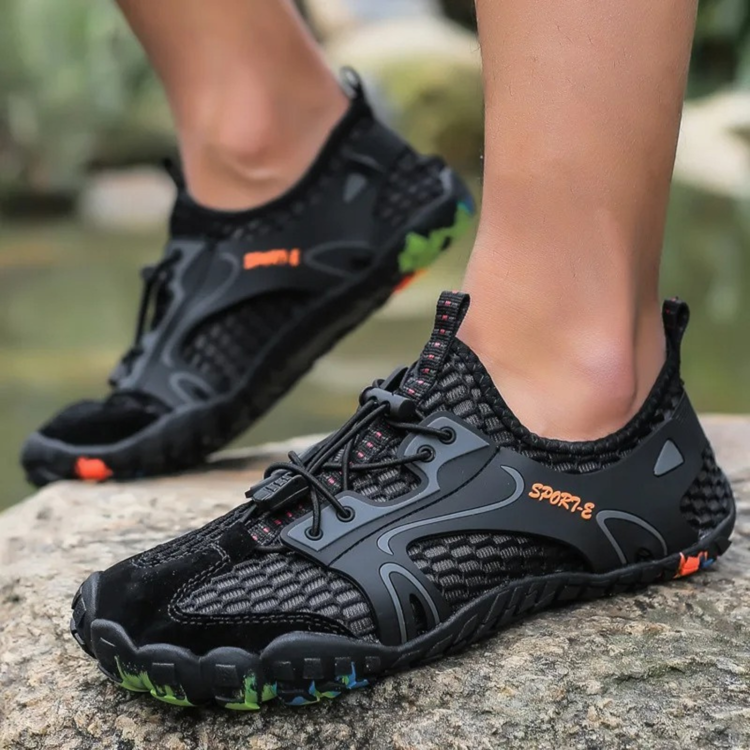 Person wearing black water shoes with rainbow sole, standing on rocky terrain.