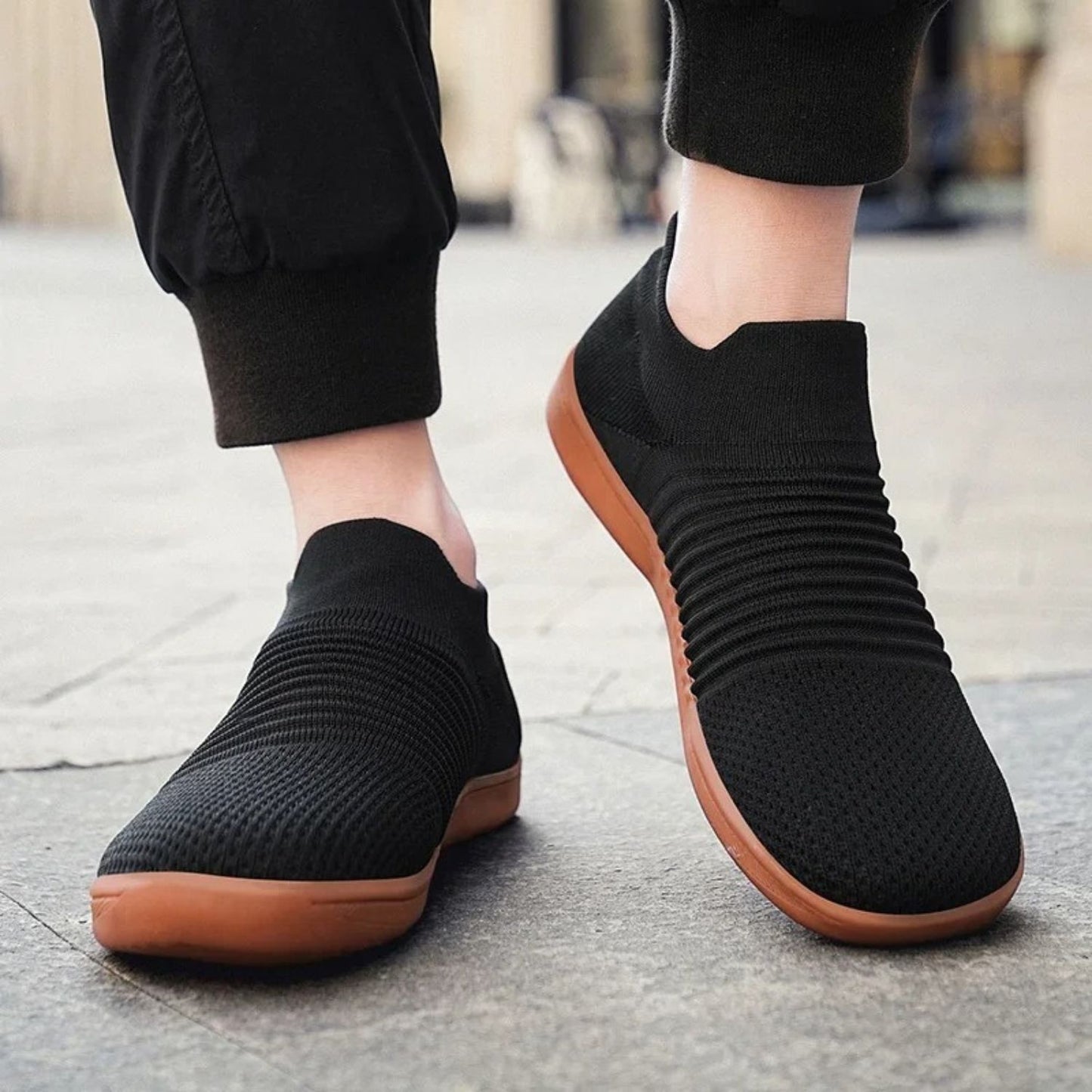 Black knit slip-on sneakers with brown soles worn with cuffed jogger pants.