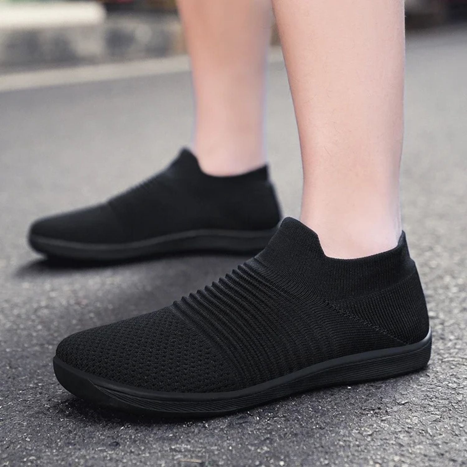 Person wearing black slip-on sneakers with ribbed texture, walking on pavement.