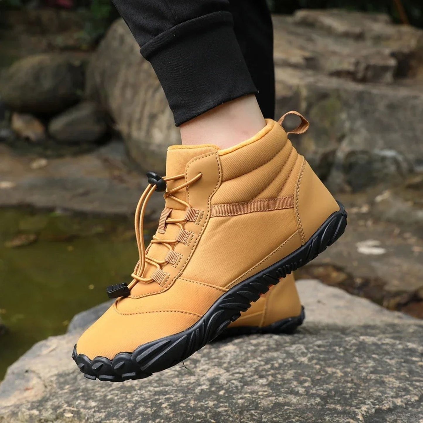 Person wearing brown hiking boots with black and orange soles, standing on rocks.